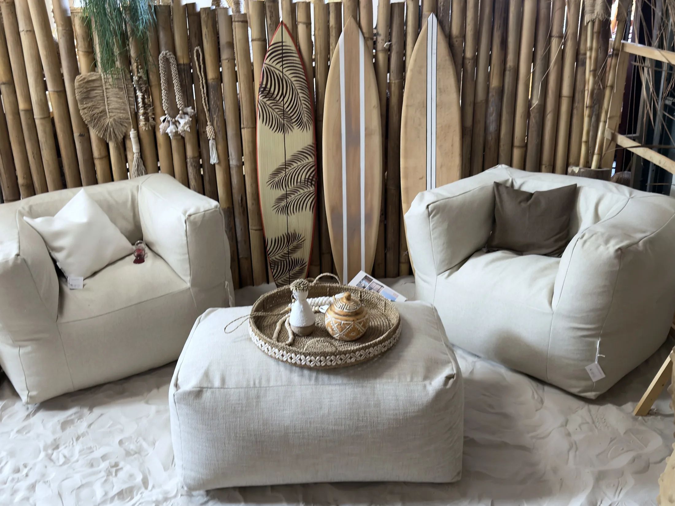 Twee linnen loungefauteuils met een poef en een rieten dienblad op de zandvloer bij CoCo homeliving, met surfplanken tegen een bamboewand