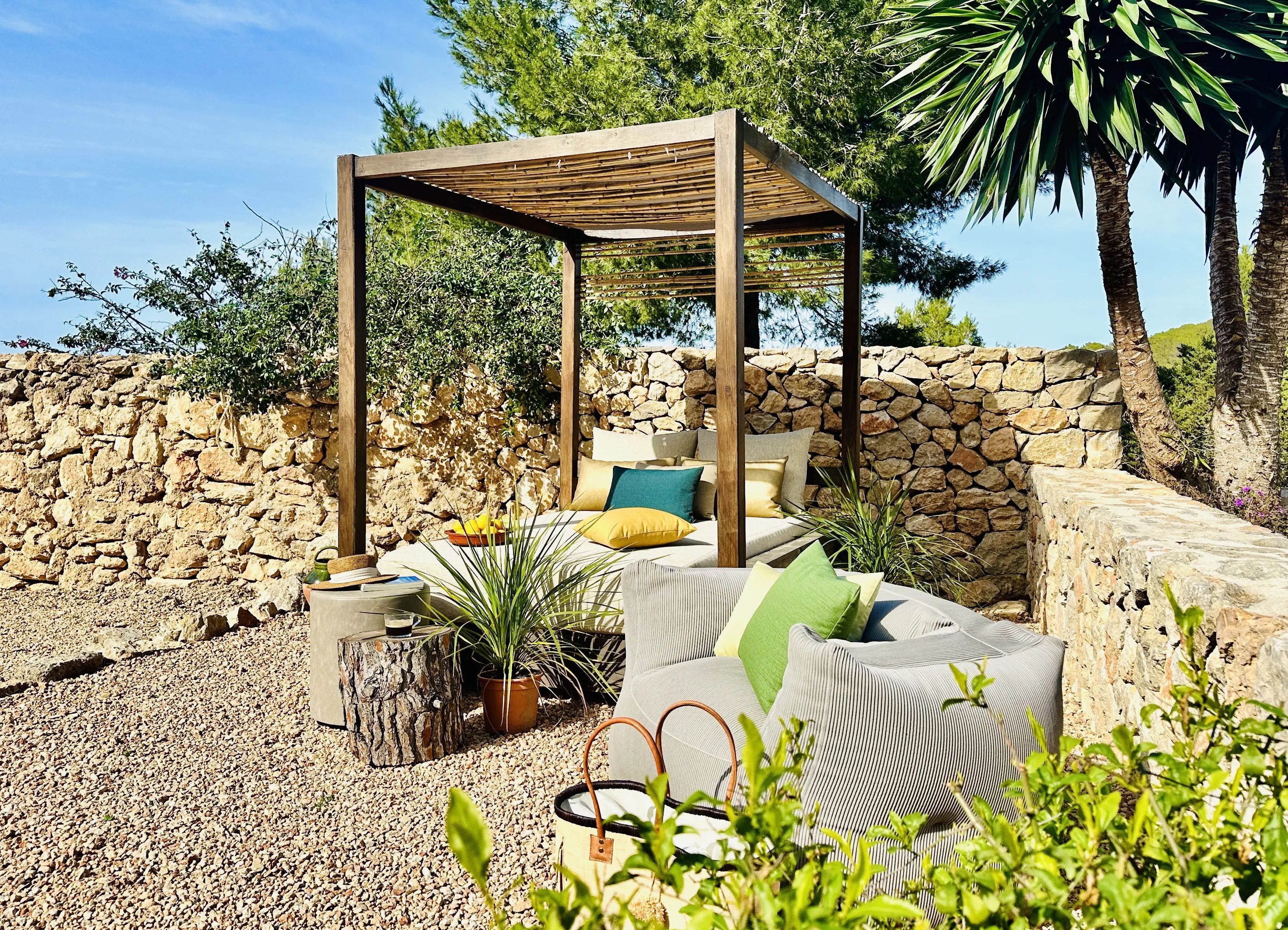 Houten pergola met bamboedak boven een daybed met kussens, en een lichtgrijze loungestoel op een grindterras langs een natuurstenen muur